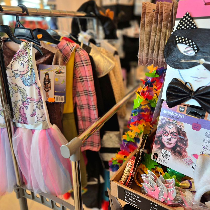 Ein Verkaufsregal zeigt eine Auswahl an Kostümen, darunter ein Einhorn-Thema-Kleid und eine pink karierte Jacke. In der Nähe befindet sich eine Box mit Halloween-Dekorationen, darunter ein Make-up-Kit, Leis und Pins.