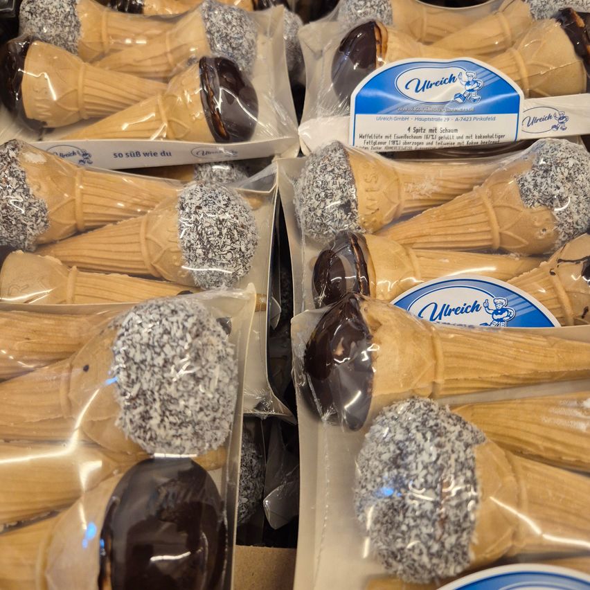 Verpackte, mit Schokolade überzogene Eiswaffeln mit Streuseln sind in durchsichtigen Plastiktüten ausgestellt. Die Kegel sind in dunkle Schokolade getaucht und haben Toppings wie Streusel und Schokoladenflakes. Der Markenname Ulrich ist auf dem Etikett sichtbar.