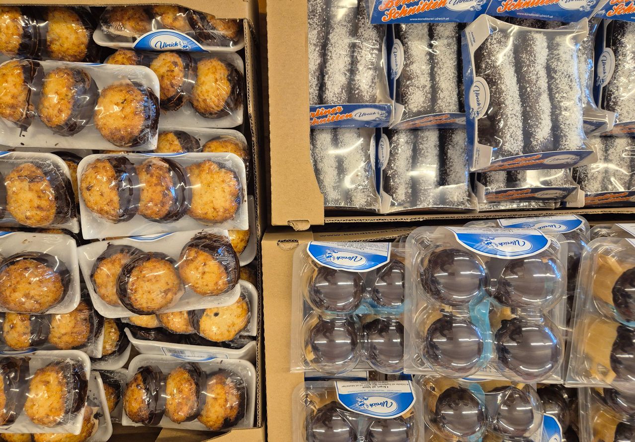 Verschiedene Schachteln mit Gebäck sind ausgestellt, darunter glasierte in transparenter Verpackung und andere in blauen Schachteln. Einige sind in braunen Kartons, andere in durchsichtigen Plastikbehältern.