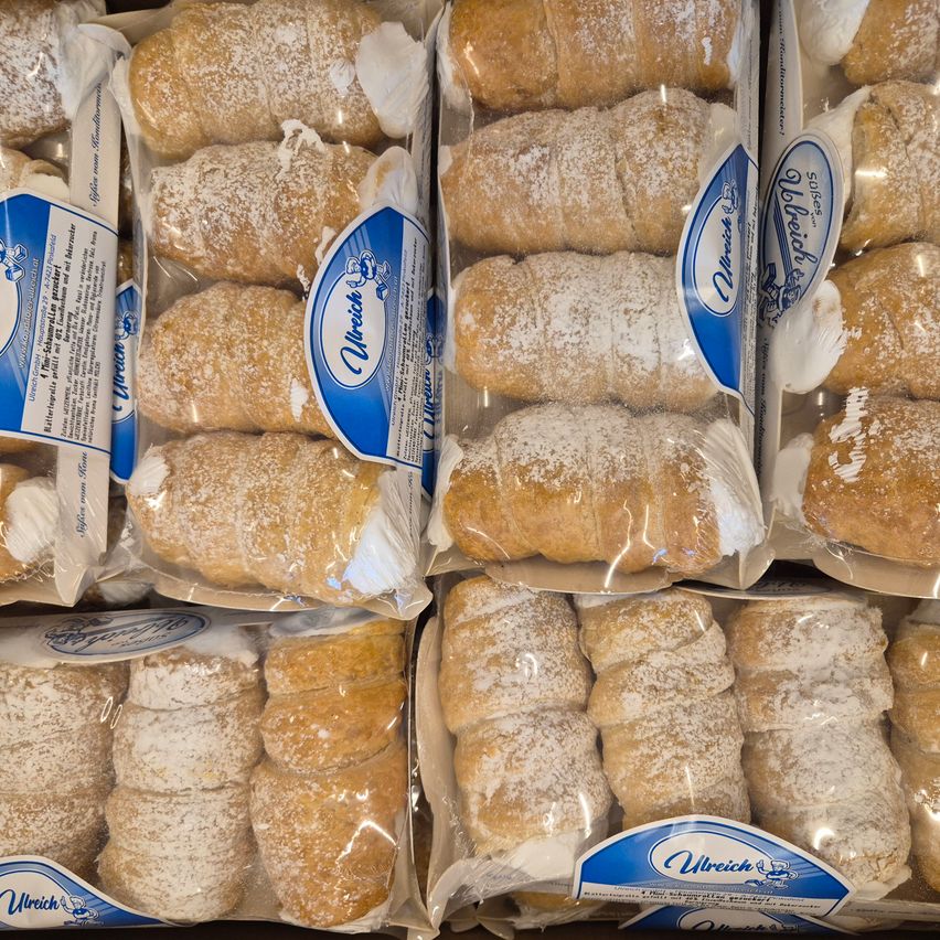 Mehrere Schachteln mit Gebäck werden gezeigt, wobei jede Schachtel mehrere Gebäckstücke enthält. Die Gebäckstücke sind spiralförmig gerollt und mit Puderzucker bestreut. Die Schachteln sind mit dem Markennamen 'Ulreich' beschriftet und haben blaue und weiße Etiketten.