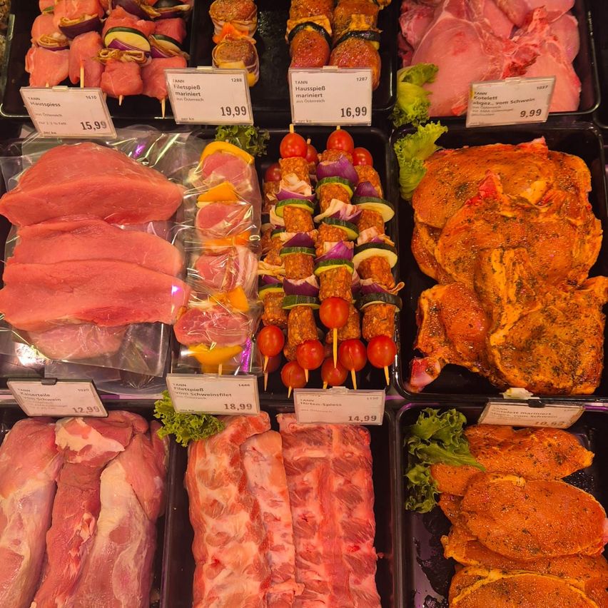 Eine Auswahl an rohem und gekochtem Fleisch, das in einem Supermarkt ausgestellt ist. Artikel umfassen Schweinekoteletts, marinierte Spieße und gewürztes Hähnchen. Die Preise sind auf Preisschildern angegeben.