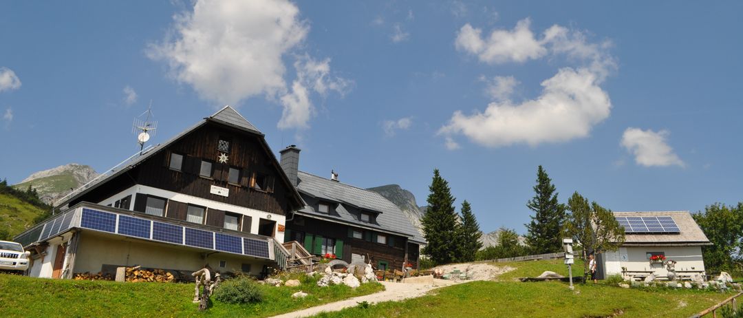 Ein Chalet mit Solarpanelen auf dem Dach, umgeben von saftigem grünem Gras und Bäumen, unter einem blauen Himmel mit Wolken.