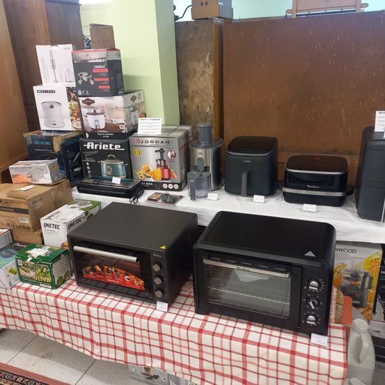 Ein Tisch mit verschiedenen Küchengeräten und Gadgets zur Schau, darunter Toaster-Öfen, Entsafter und Reiskocher. Der Tisch ist mit einer rot-weiß karierten Tischdecke bedeckt.