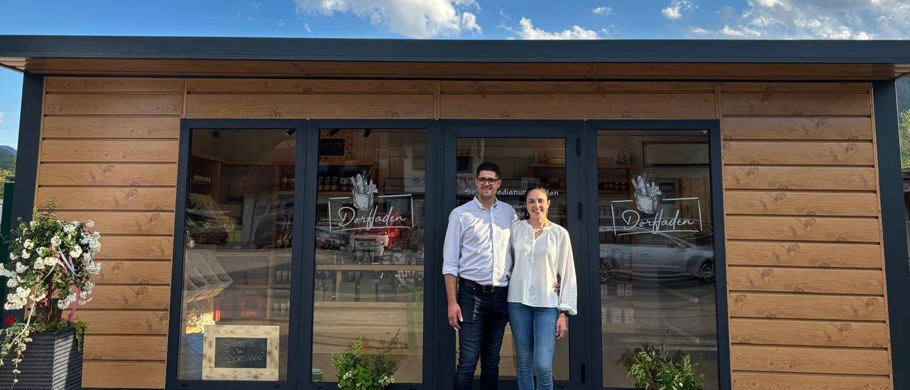 Zwei Personen stehen vor einem Geschäft namens Dorfladen mit blauem Himmel und Wolken im Hintergrund. Das Geschäft hat Glastüren und Fenster, und es gibt Pflanzen und Blumen draußen.