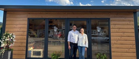 Two people stand in front of a shop named Dorfladen with a blue sky and clouds in the background. The shop has glass doors and windows, and there are plants and flowers outside.