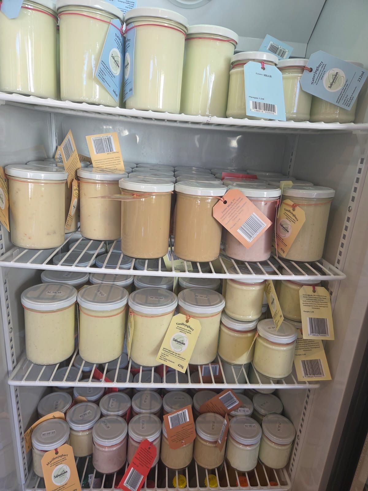 A refrigerator shelf filled with jars of different colored creams and sauces, labeled with tags, stored on white shelves.