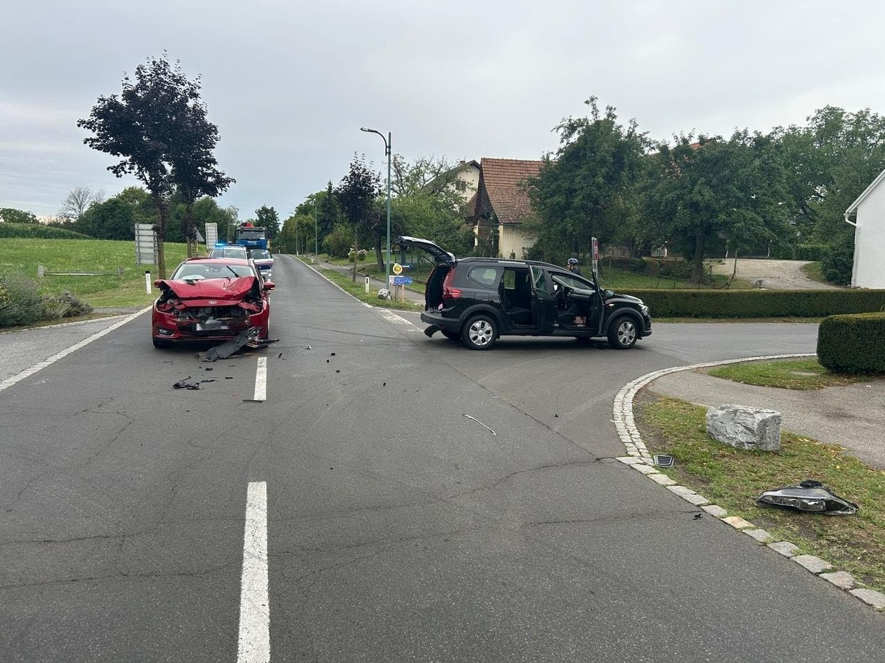 Ein rotes Auto wurde auf einer Straße beschädigt, in der Nähe ein schwarzer SUV. Die Tür des SUV ist offen. Im Hintergrund befinden sich Bäume und ein Haus.