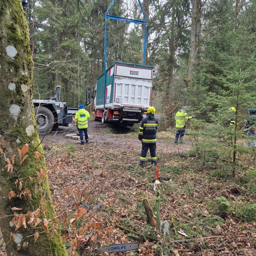 Feuerwehrleute in Schutzkleidung arbeiten in einem Wald. Ein Lkw wird von einem Kran angehoben. Eine Kettensäge liegt auf dem Boden.
