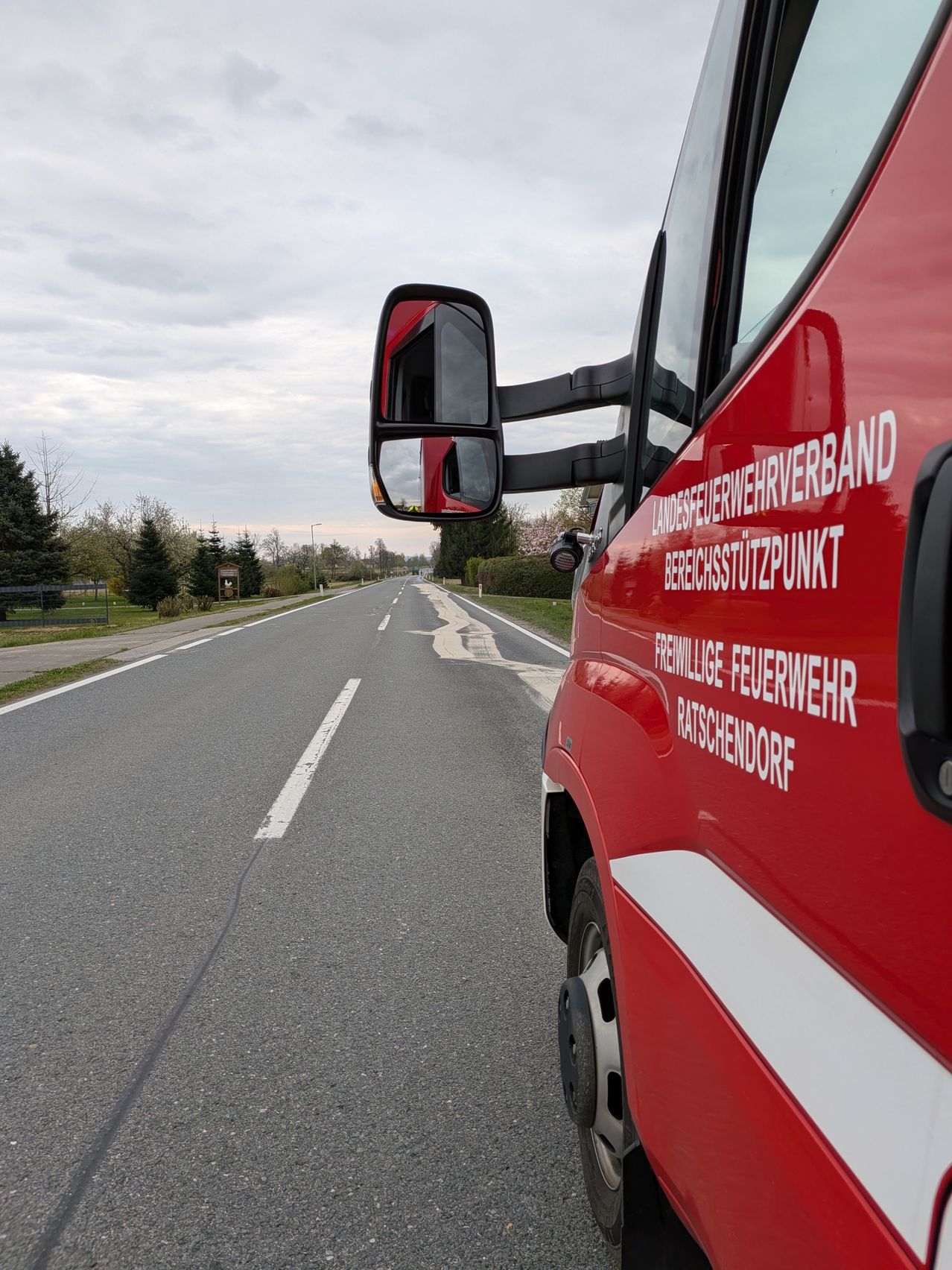 A red fire truck is parked on the side of a road with white markings. Trees line the road, and a cloudy sky is visible above.