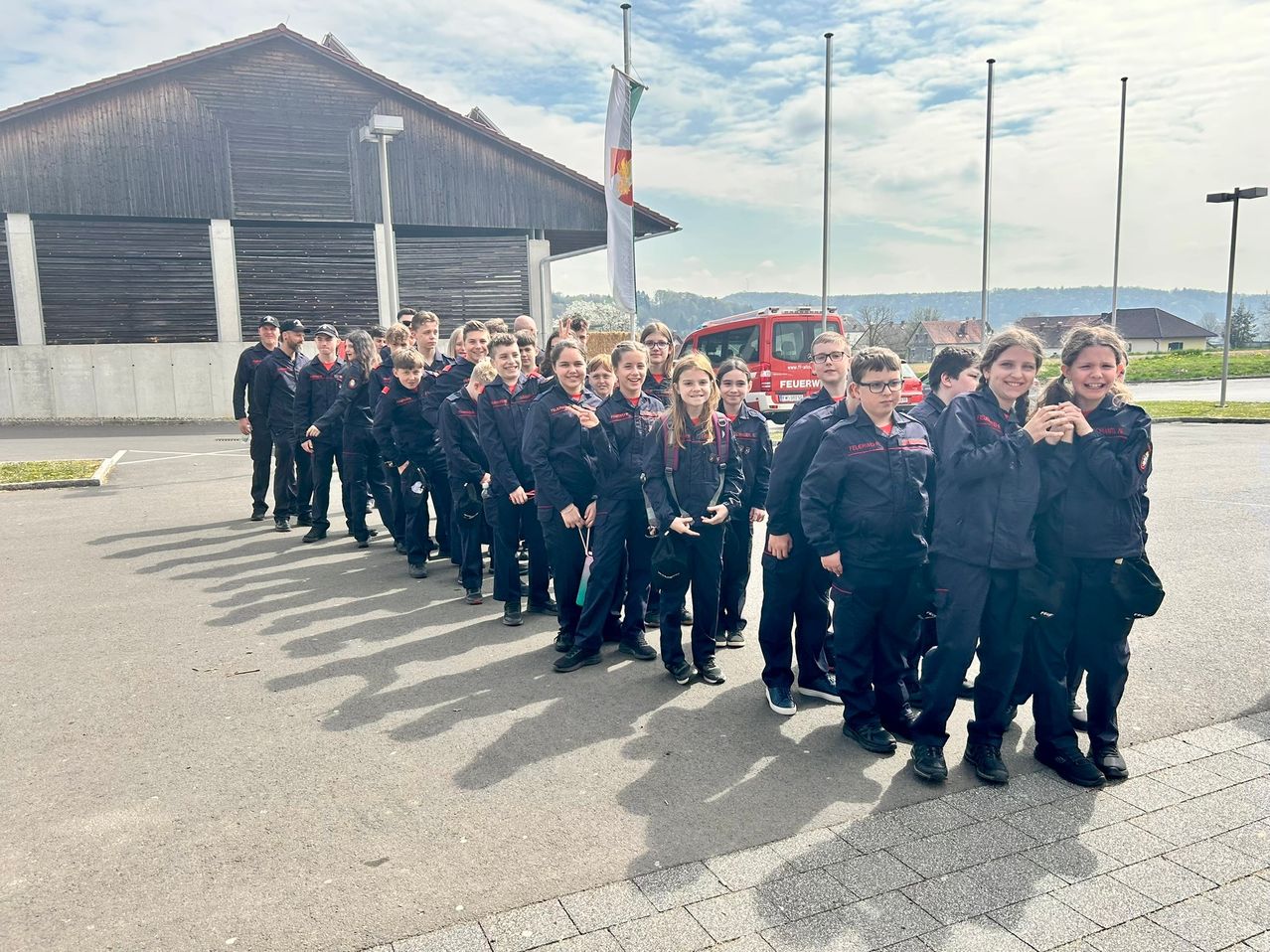 A group of people dressed in uniform are standing in a row, possibly for a photo. They are in front of a building with a flag and a fire truck.