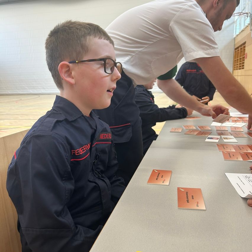 A boy with glasses sits at a table with cards while a man in a white shirt helps him. Other people are around them.
