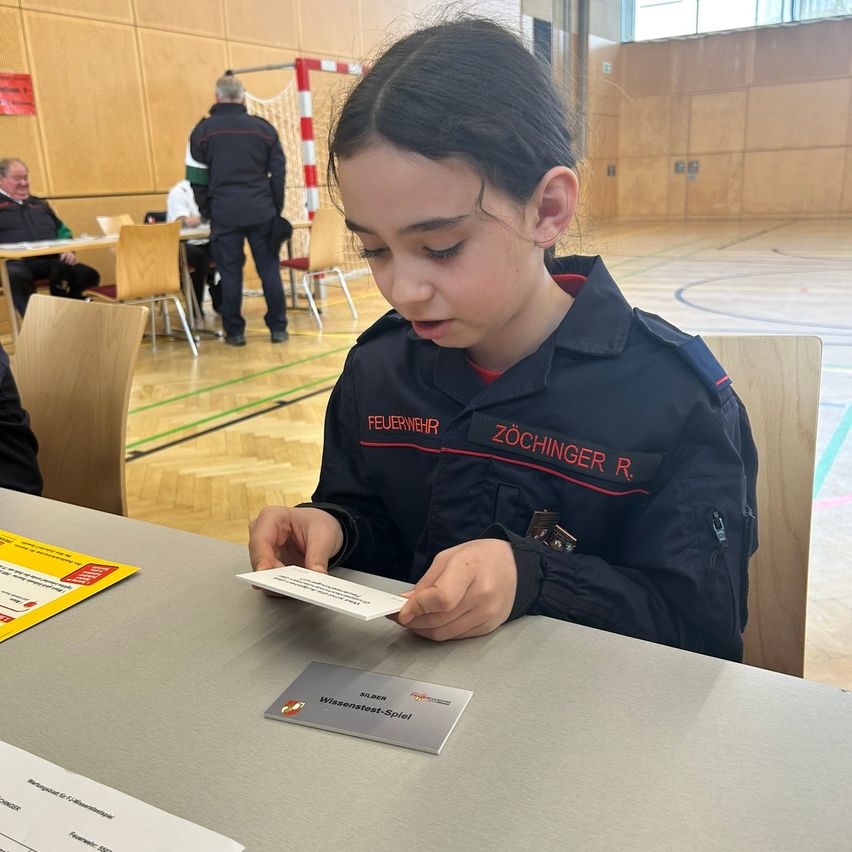 A girl in a uniform reads a card in a room with other people and tables.