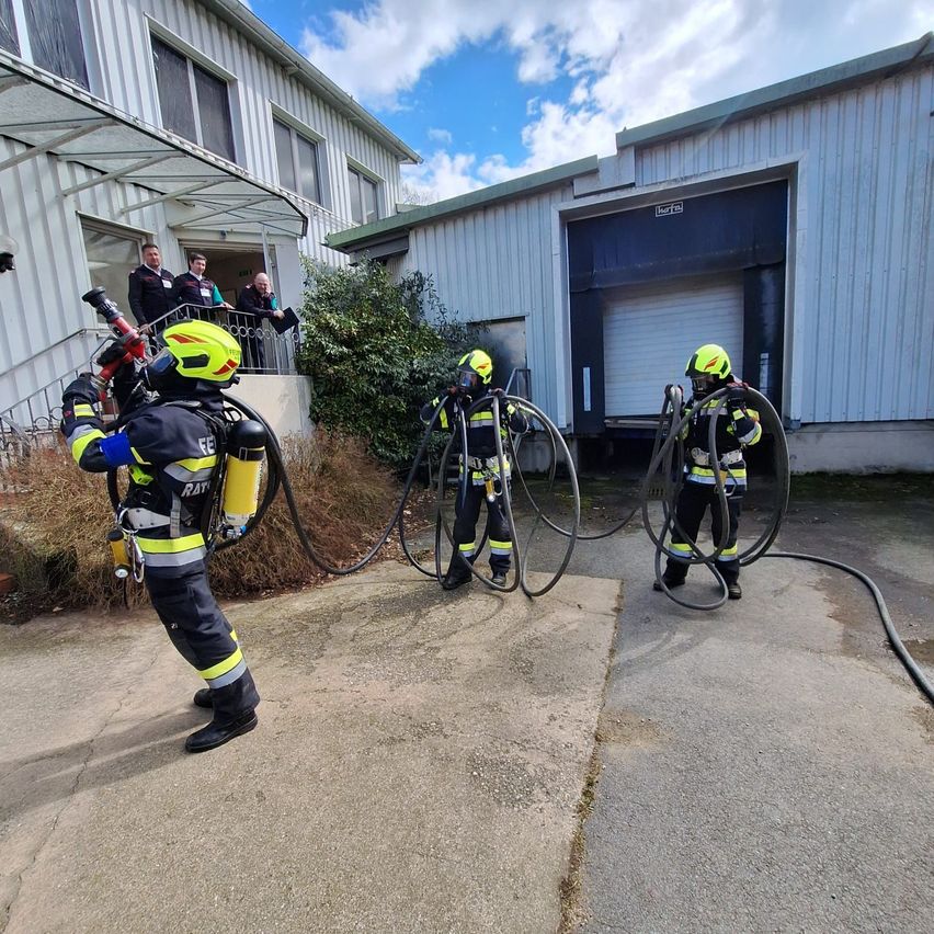 Eine Gruppe von Feuerwehrleuten übt das Benutzen von Feuerwehrschläuchen vor einem Gebäude. Ein Feuerwehrmann hält einen Schlauch, während die anderen hinter ihm stehen.