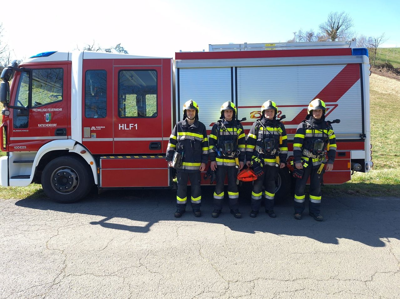 Vier Feuerwehrleute stehen vor einem roten Feuerwehrauto mit der Aufschrift HLF1, jeder trägt ein gelb-schwarzes Uniform. Das Auto hat Glasfenster und eine Tür. Bäume sind im Hintergrund zu sehen.