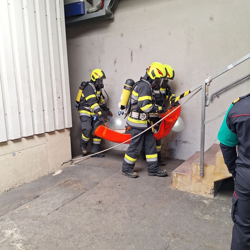 Drei Feuerwehrleute in voller Ausrüstung, die eine rote Trage halten, verlassen ein Gebäude. Einer steht auf der Treppe, ein anderer auf dem Betonboden.
