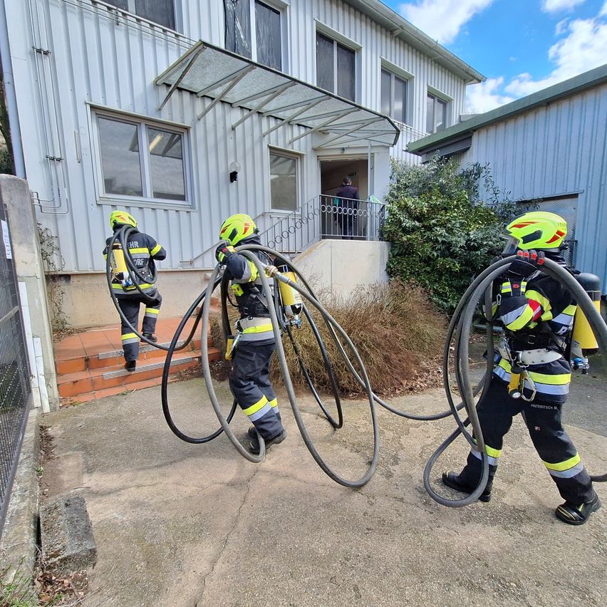 Drei Feuerwehrleute tragen Schläuche in der Nähe eines Gebäudes mit Balkon und Treppe, wobei eine Person eintritt.