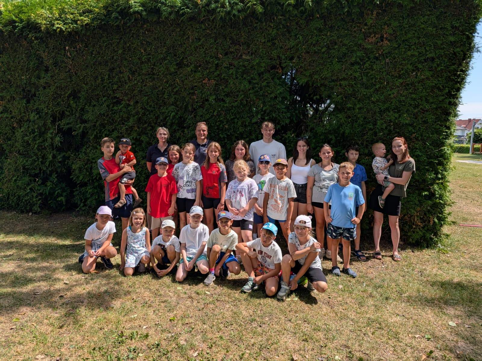 Kinder und Betreuer stehen und sitzen vor einer Hecke in einem Park.