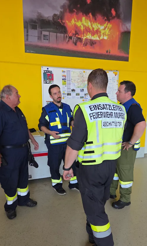 Vier Feuerwehrleute stehen vor einer gelben Wand mit einem Feuerposter und diskutieren etwas.