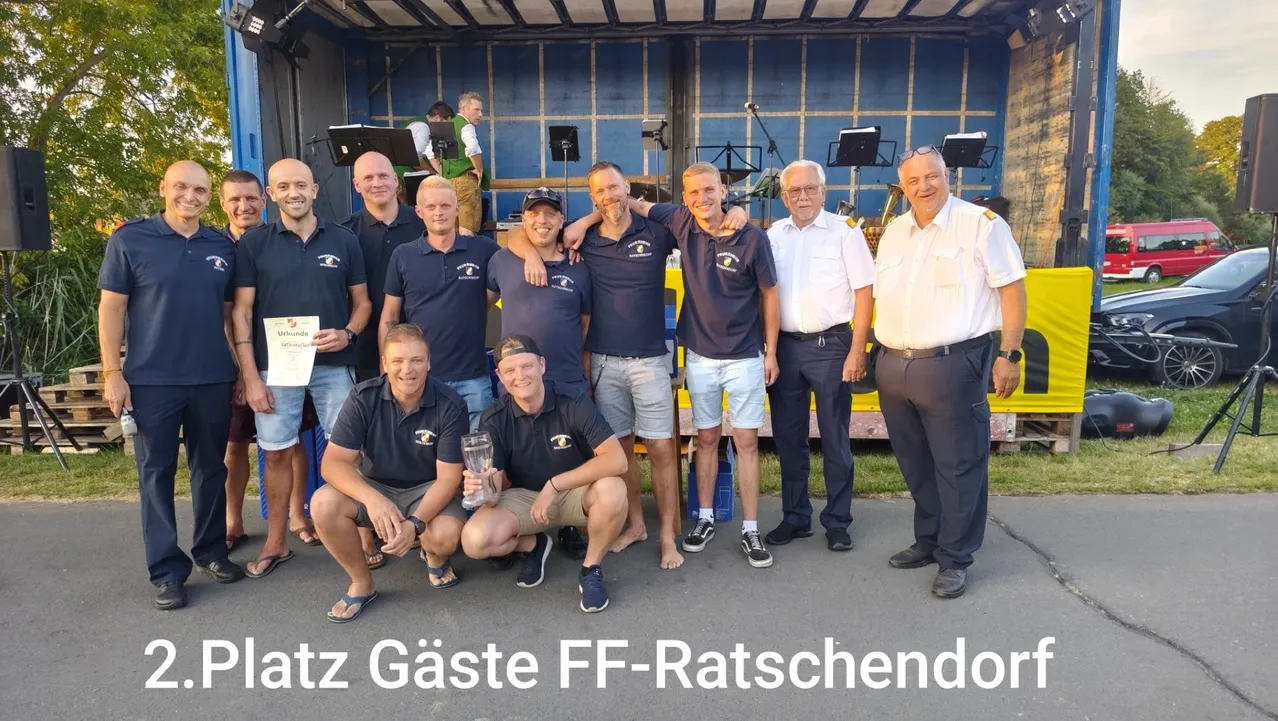 Eine Gruppe von Männern in blauen Poloshirts und Shorts posiert für ein Foto mit einer Trophäe. Sie stehen vor einem blauen Hintergrund mit Musikinstrumenten und einem Schild, das 'Ratz Gaste FF-Ratschendo' besagt.