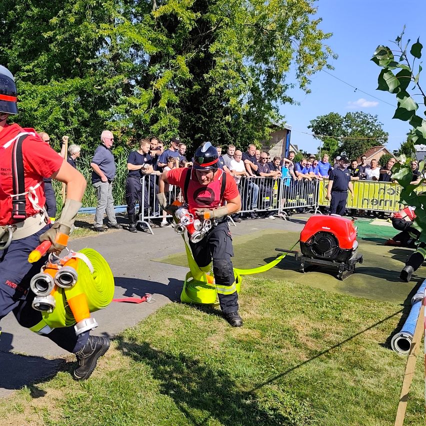 Zwei Feuerwehrleute in roten Uniformen führen eine Demonstration vor einer Menge durch. Einer hält eine Feuerwehrspritze, während der andere auf einer grünen Plattform mit einem Feuerlöscher steht.
