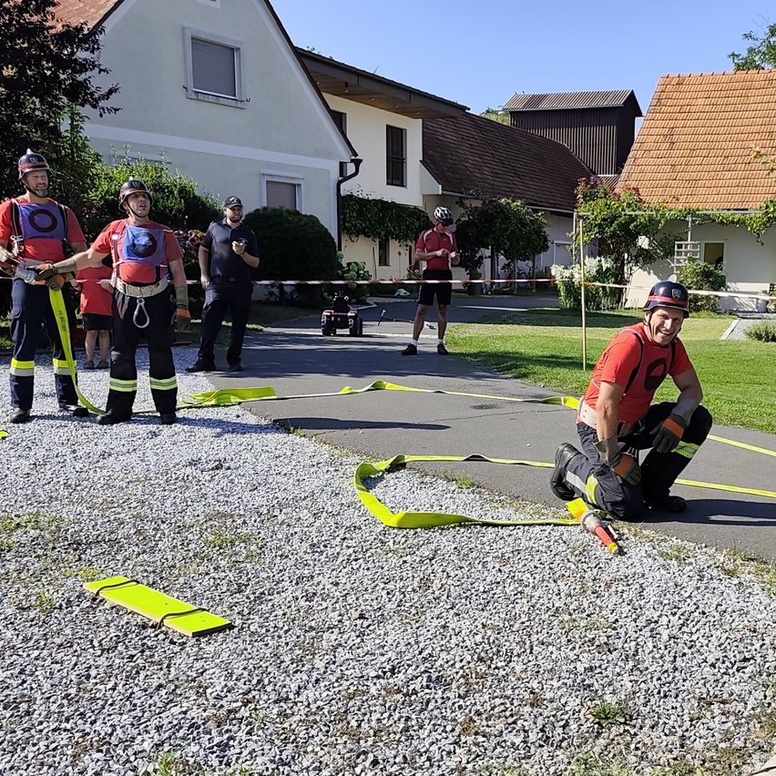 Eine Gruppe von Feuerwehrleuten in voller Ausrüstung übt auf einem Kiesweg, einer kniet und einer hält ein Seil. Hinter ihnen sind Häuser und Grünflächen zu sehen.