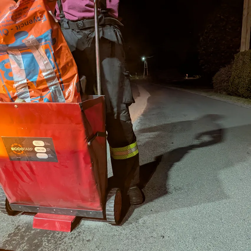 Eine Person in reflektierender Kleidung schiebt einen roten Wagen mit einem Sack Dünger bei Nacht auf einer Straße.