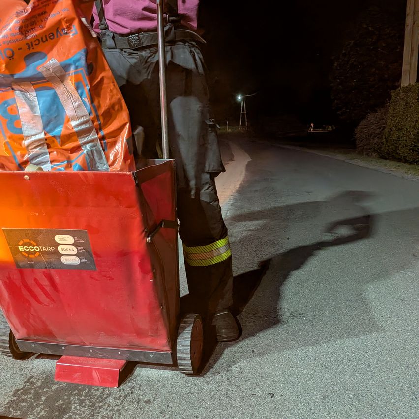 Eine Person in reflektierender Kleidung schiebt einen roten Wagen mit einem Sack Dünger bei Nacht auf einer Straße.
