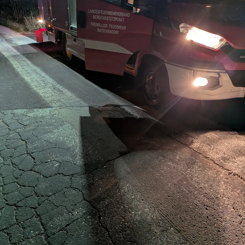 Ein roter Feuerwehrwagen ist am Straßenrand geparkt, mit eingeschalteten Scheinwerfern, die einen beschädigten Abschnitt des Bürgersteigs beleuchten.