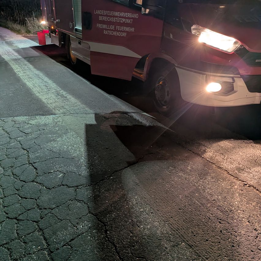 Ein roter Feuerwehrwagen ist am Straßenrand geparkt, mit eingeschalteten Scheinwerfern, die einen beschädigten Abschnitt des Bürgersteigs beleuchten.