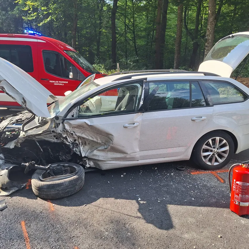 Ein weißer Wagen mit offenem Motorhaube und beschädigtem vorderen Stoßfänger liegt am Straßenrand, mit einem Krankenwagen in der Nähe.