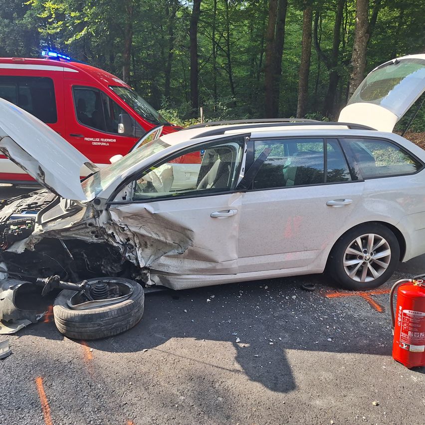 A white car with its hood open and front bumper damaged lies on the side of a road, with an ambulance nearby.