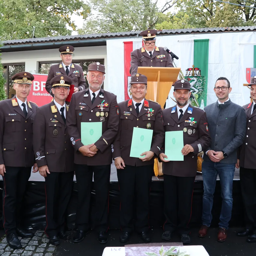 Eine Gruppe von Männern in Militäruniformen steht auf einer Bühne und hält Urkunden in den Händen, während ein Mann in einem Anzug neben ihnen steht. Hinter ihnen ist ein Podium mit einem Mikrofon zu sehen, und eine Flagge ist an der Wand angebracht.
