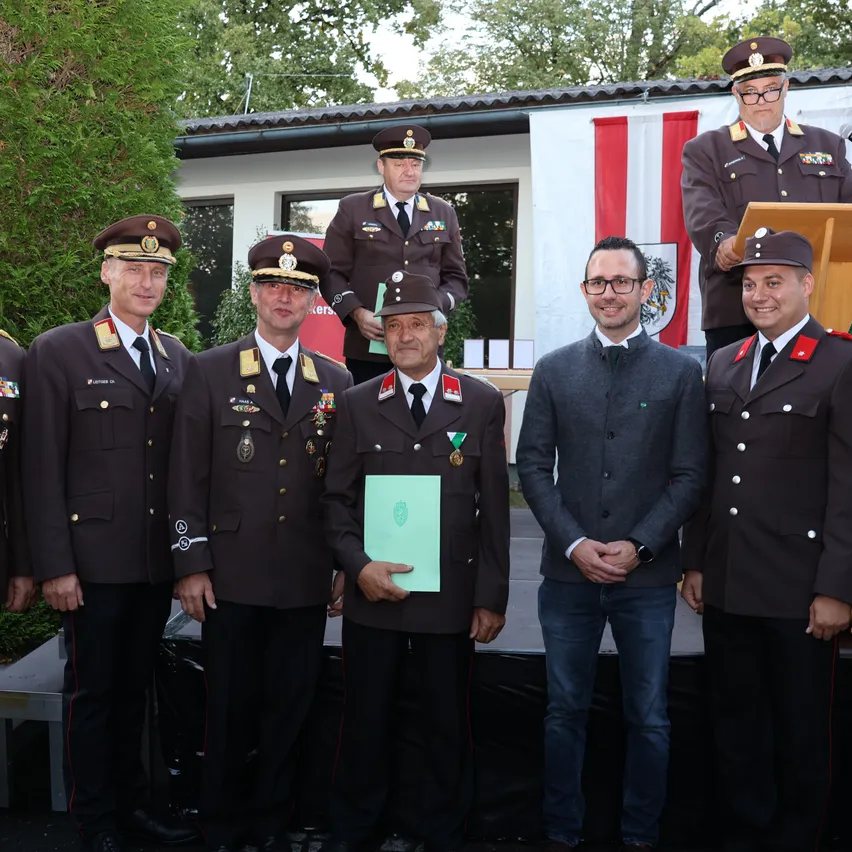 Eine Gruppe von Männern in Militäruniformen steht zusammen auf einer Bühne, einer hält ein grünes Dokument. Dahinter ist eine Fahne mit einem Banner angebracht.