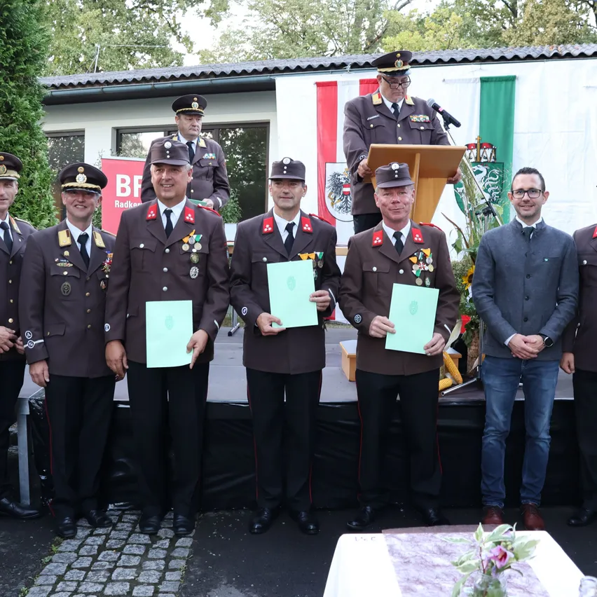 Eine Gruppe von Männern in Militäruniformen steht auf einer Bühne und hält Urkunden. Ein Mann in einem Anzug steht neben ihnen. Dahinter ist ein Podium mit Mikrofon und Flaggen zu sehen.
