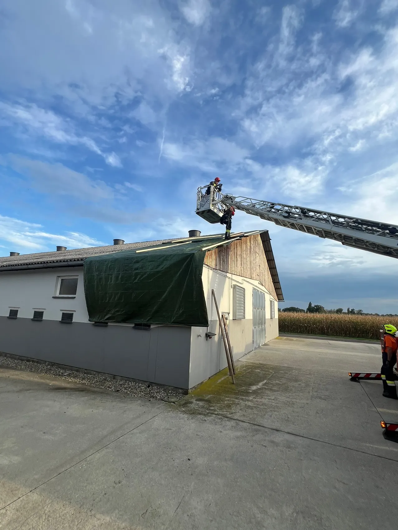 Feuerwehrleute auf einer Leiter sichern eine grüne Plane über dem Dach eines Gebäudes. Das Gebäude hat einen hölzernen Teil und ist von einem Kiesbereich umgeben. Der Himmel ist teilweise bewölkt.