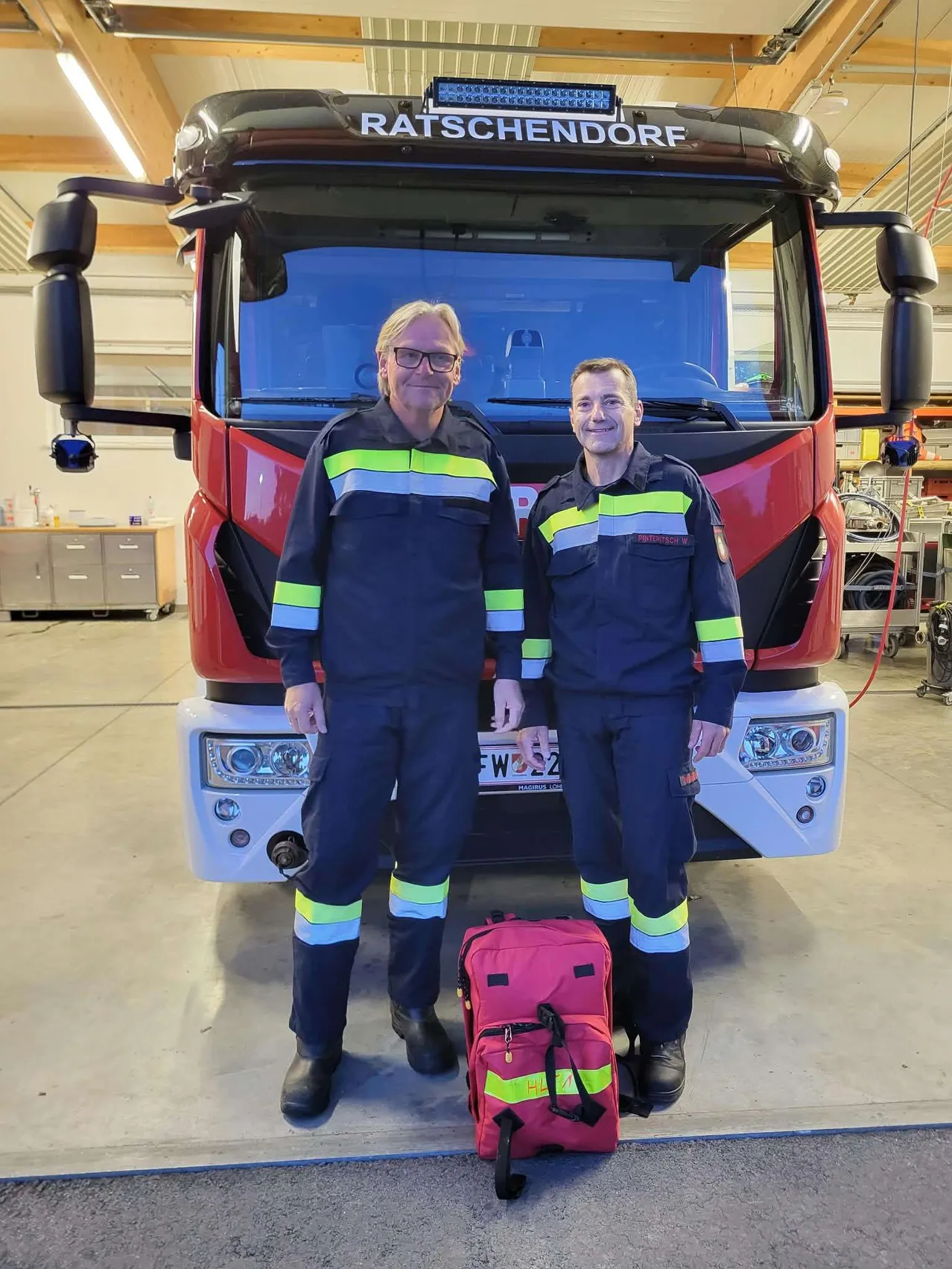 Zwei Feuerwehrleute stehen vor einem roten Feuerwehrauto und lächeln für ein Foto.