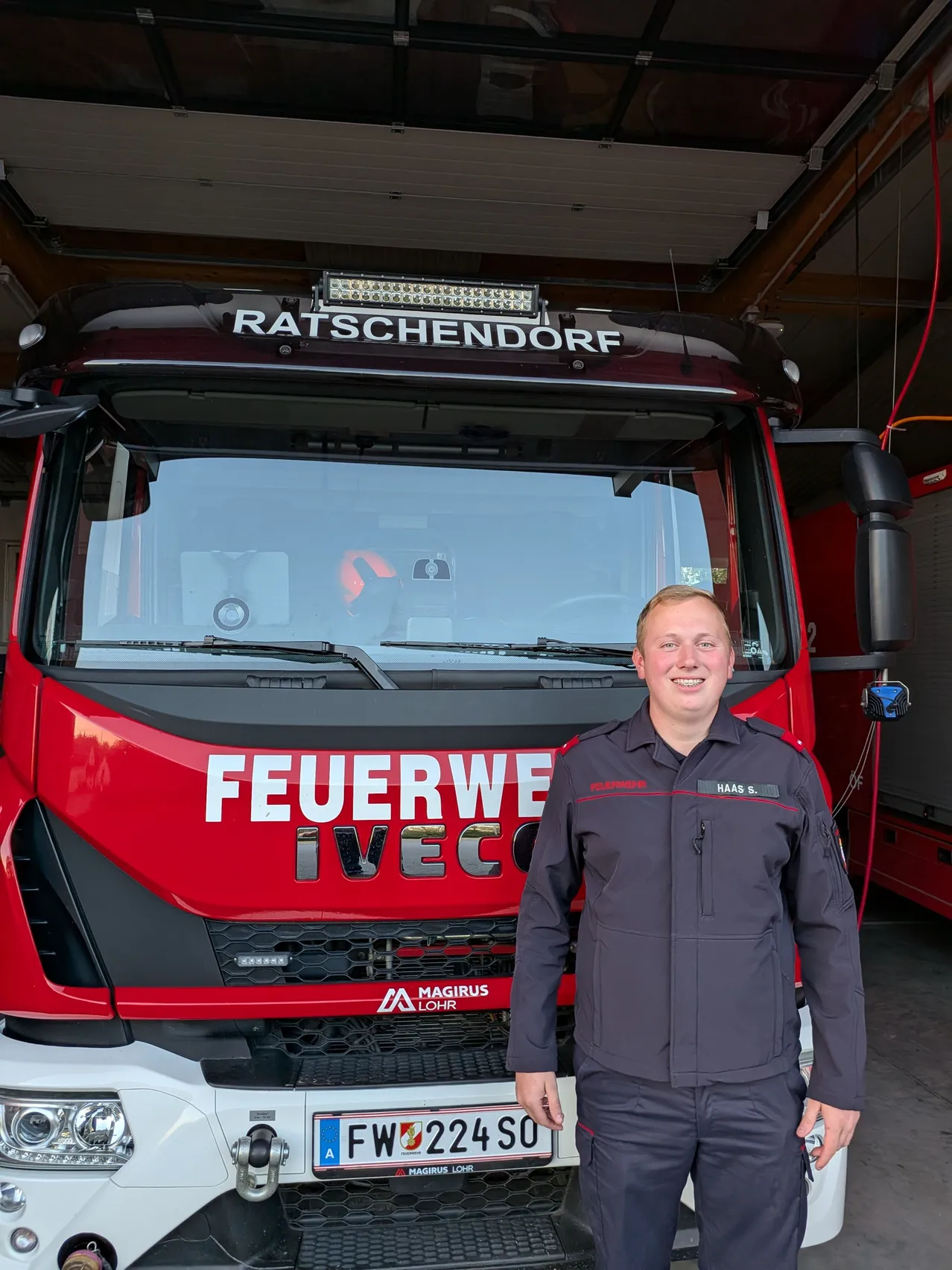 Ein Feuerwehrmann steht vor einem roten Iveco-Löschfahrzeug in einer Garage. Das Fahrzeug hat die Aufschrift 'FEUERWEHR' und den Ortsnamen 'RATSCHENDORF' auf der Front. Der Feuerwehrmann trägt eine Uniform und lächelt.