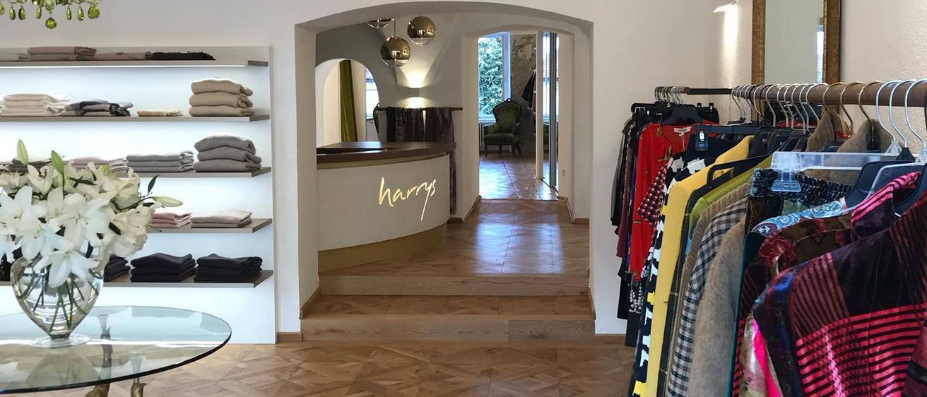 Ein Boutique-Interieur mit Holzböden, weißen Wänden und einer Kristallleuchte. Ein Glastisch mit einer Vase mit weißen Blumen. Kleidung hängt an Kleiderbügeln, und ein Schild steht 'harry'.