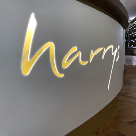 Eine gebogene weiße Wand mit 'Harrys' in gelben kursiven Buchstaben. Ein hölzerner Boden mit Parkett-Design. Dahinter eine Glastür mit einem Regal im Inneren.