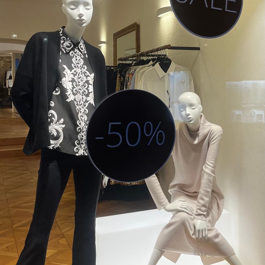 Zwei Mannequins in Kleidung sind in einem Geschäft. Ein Rabattschild zeigt -50%. Das Mannequin rechts trägt ein weißes Kleid.