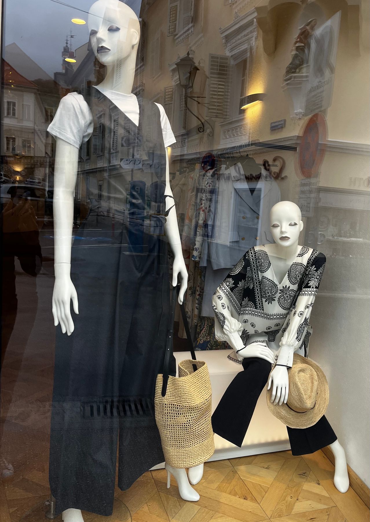 A clothing store display with mannequins wearing black dresses and hats. The left mannequin has a woven bag on its shoulder.