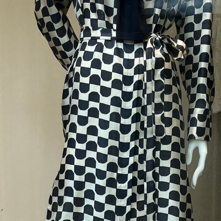 A mannequin displays a long, black and white checkered dress with long sleeves and a waist tie.