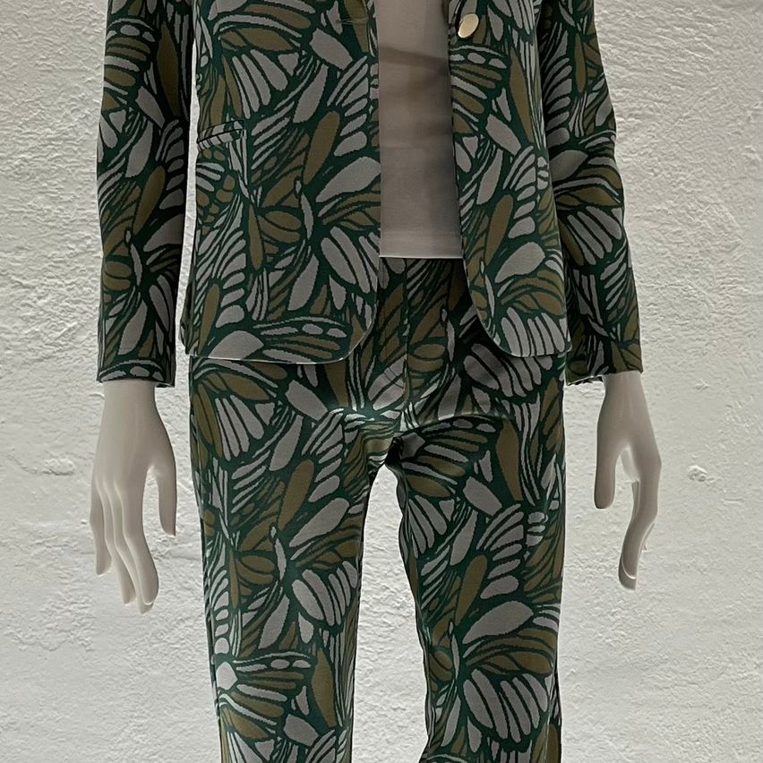 Eine Mannequin in einem grünen gemusterten Blazer und dazu passenden Hosen vor einer weißen Wand.