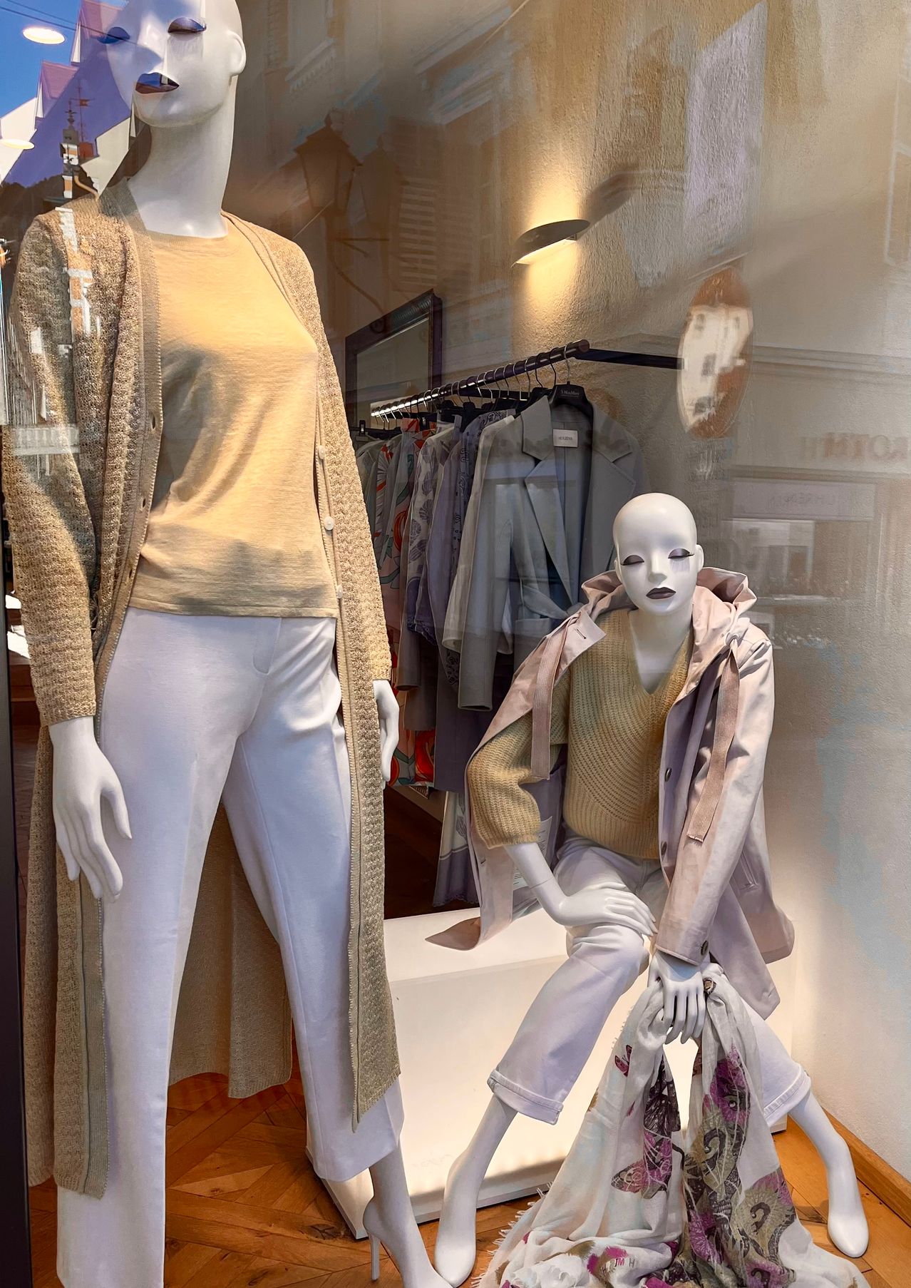 Eine Puppe in einem beigen Cardigan und weißen Hosen steht neben einer anderen Puppe in einem beigen Pullover und einer beigen Jacke. Dahinter hängen Kleider an einem Bügel.