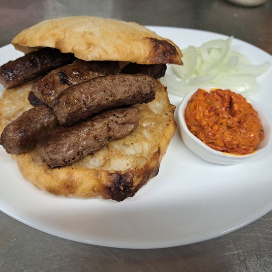 Ein Teller mit einem Fleischbrötchen, in Stücke geschnitten, auf einem Brötchen mit gegrillten Spuren, begleitet von geschnittenen Zwiebeln und einer roten Sauce.