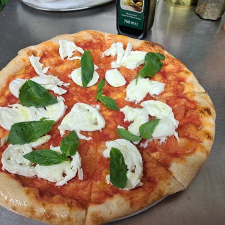 Eine runde Pizza, die mit frischen Basilikumblättern und Mozzarellakäse belegt ist, steht auf einem Metalltisch. Im Hintergrund befindet sich eine Flasche Olivenöl.