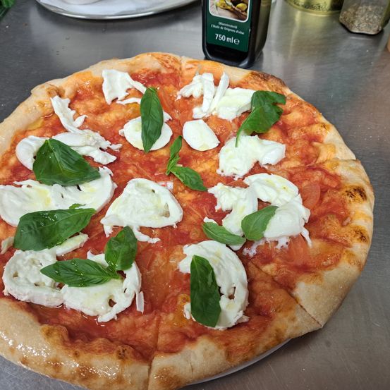Eine runde Pizza, die mit frischen Basilikumblättern und Mozzarellakäse belegt ist, steht auf einem Metalltisch. Im Hintergrund befindet sich eine Flasche Olivenöl.