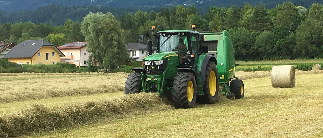 Ein grüner Traktor erntet Feldfrüchte auf einem Feld. Eine Person fährt ihn. Das Feld ist von Bäumen und Bergen umgeben.