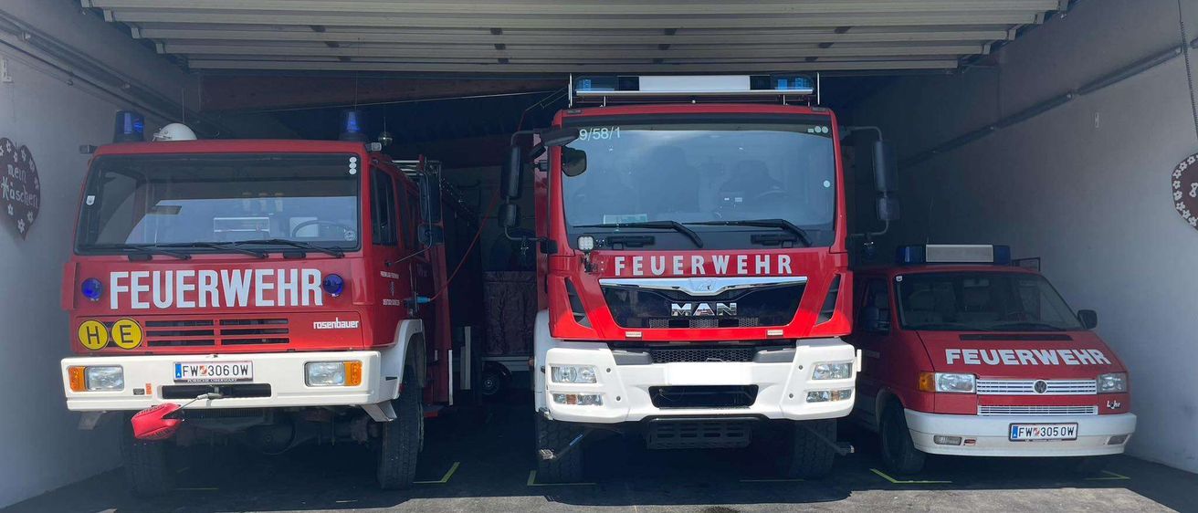 Drei Feuerwehrfahrzeuge sind in einer Garage geparkt. Das mittlere Fahrzeug hat den Schriftzug 'Feuerwehr' auf der Front. Die Fahrzeuge links und rechts sind teilweise sichtbar.