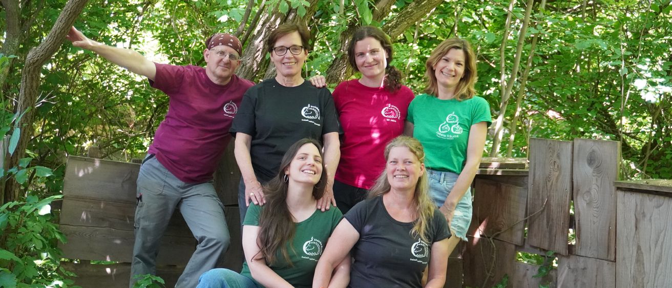 Eine Gruppe von sechs Erwachsenen, die passende T-Shirts mit Logos tragen, posiert für ein Foto in einem Wald. Sie lächeln und einige lehnen sich an einen Holzzaun.